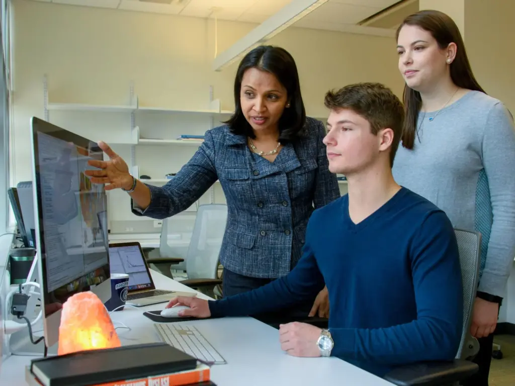 A professor pointing at a computer screen while explaining something to two students in an office setting. One student is seated, using the computer, while the other stands nearby. The background features shelves and a well-lit workspace.