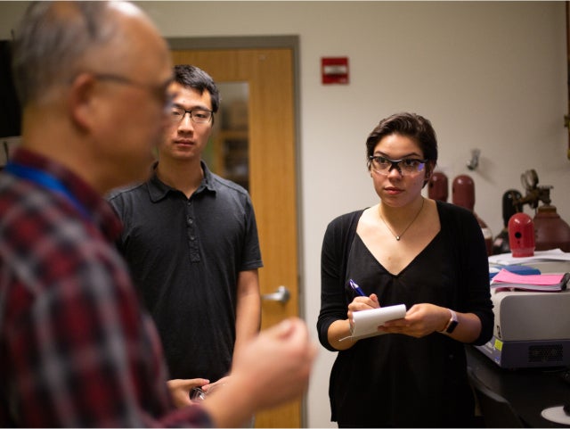 Three people are standing in a room. One person is holding a notepad and pen, appearing to take notes. The background includes a door and some lab equipment on a counter.