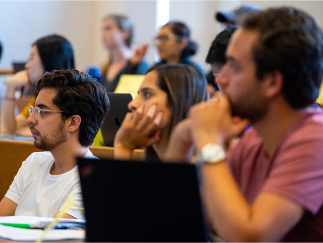 classroom setting with several students seated at desks, attentively listening and taking notes. Some students have laptops open in front of them. The room is well-lit with natural light coming through the windows. The students appear engaged in the lecture or discussion.