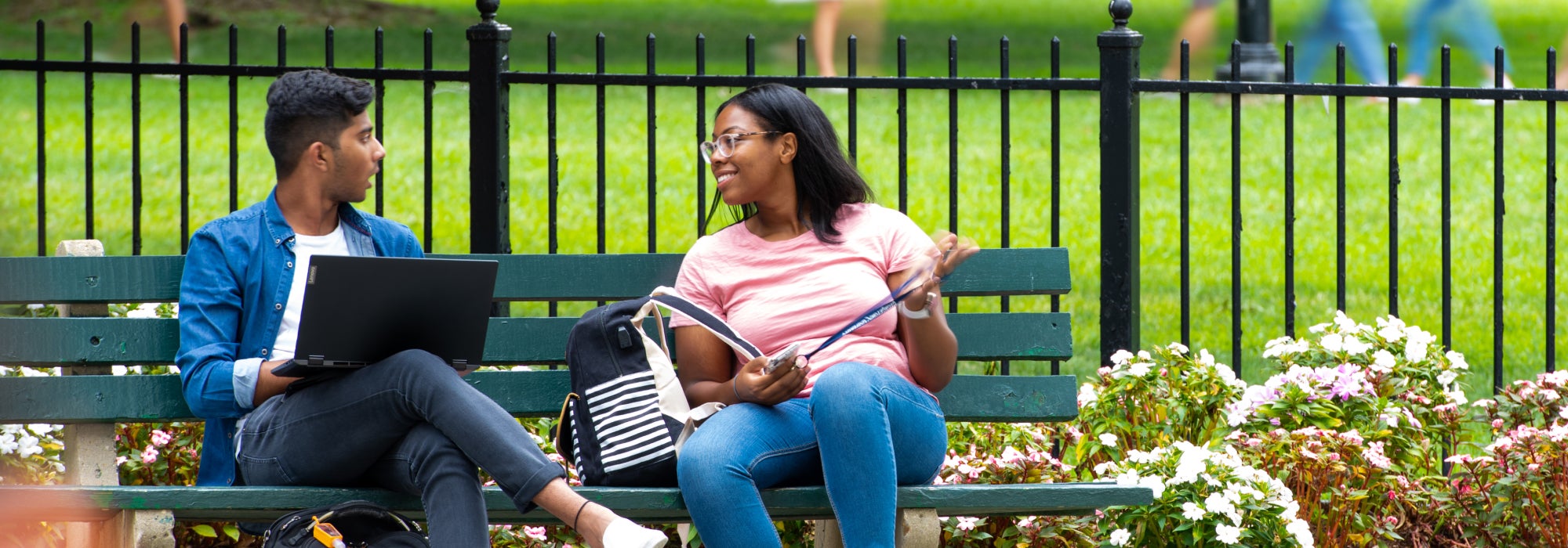 Two students sitting on a bench outdoors, engaged in conversation. One holds a laptop while the other gestures with a pen. The background features a grassy area, a black fence, and colorful flowers in the foreground.