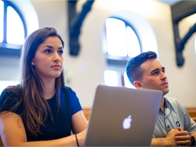 Two students attentively listening during a class session in a room with large arched windows. The student in the foreground is using a laptop, while the other sits nearby, looking focused.