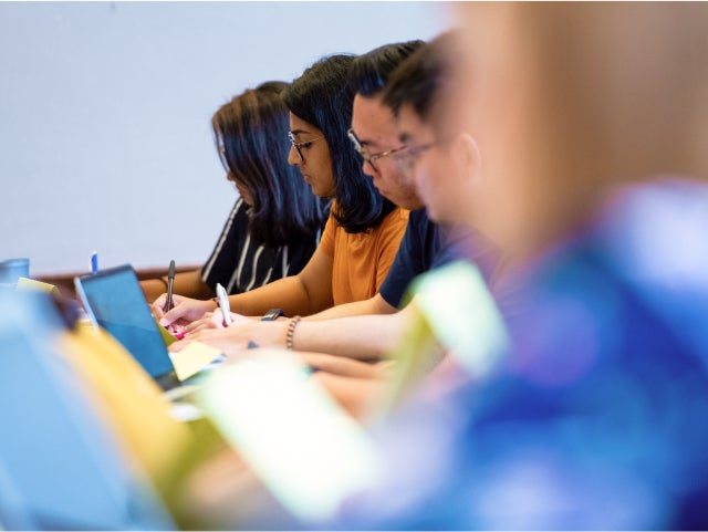 A group of people sitting at a table, focusing on their laptops and notebooks. Some individuals are writing with pens, while others are typing on their laptops. The image captures a learning or working environment, with blurred faces to maintain privacy. The background is plain, emphasizing the concentration and activity of the group.