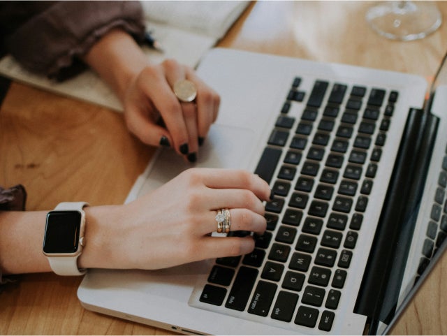 Close-up of a person working on a laptop, showing hands with jewelry and a smartwatch on a wooden desk