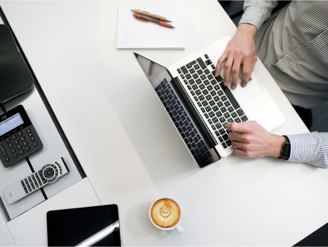 "Overhead view of a person typing on a laptop at a white desk. The desk also features a cappuccino, a notebook with pens, a tablet, and office equipment
